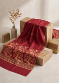 A deep red silk-like fabric with intricate gold floral and geometric patterns draped over wooden blocks, styled with a neutral beige background and a small vase of dried branches.