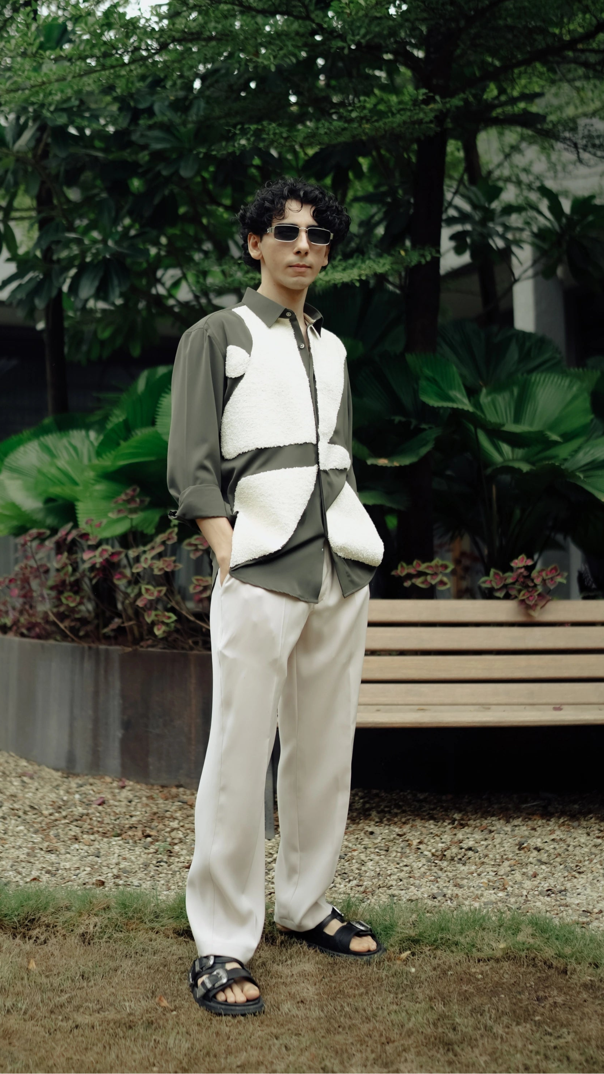 Person wearing a distinctive vest standing outdoors with greenery and a bench in the background