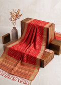 A red and gold patterned shawl with fringed edges draped over stacked wooden display blocks, with a small vase of dried branches on a light textured background.